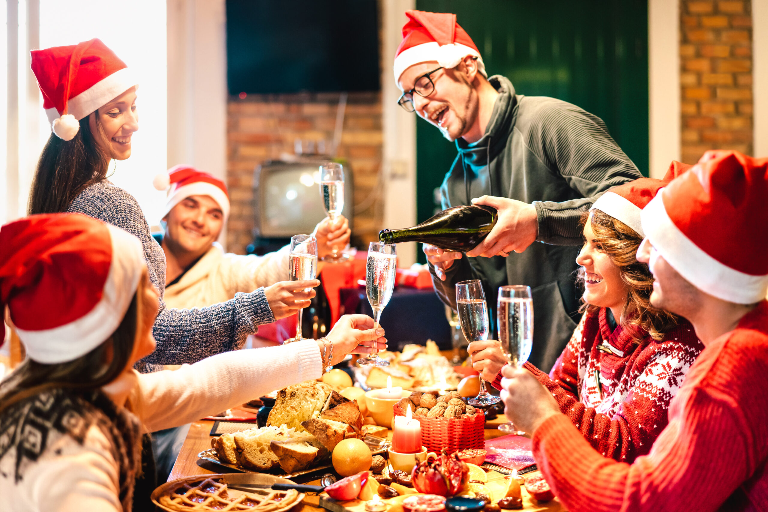 Millenial friends on santa hats celebrating Christmas with champagne and sweets food at log cabin &#8211; Winter holidays concept with young people enjoying time and having fun eating together &#8211; Warm filter